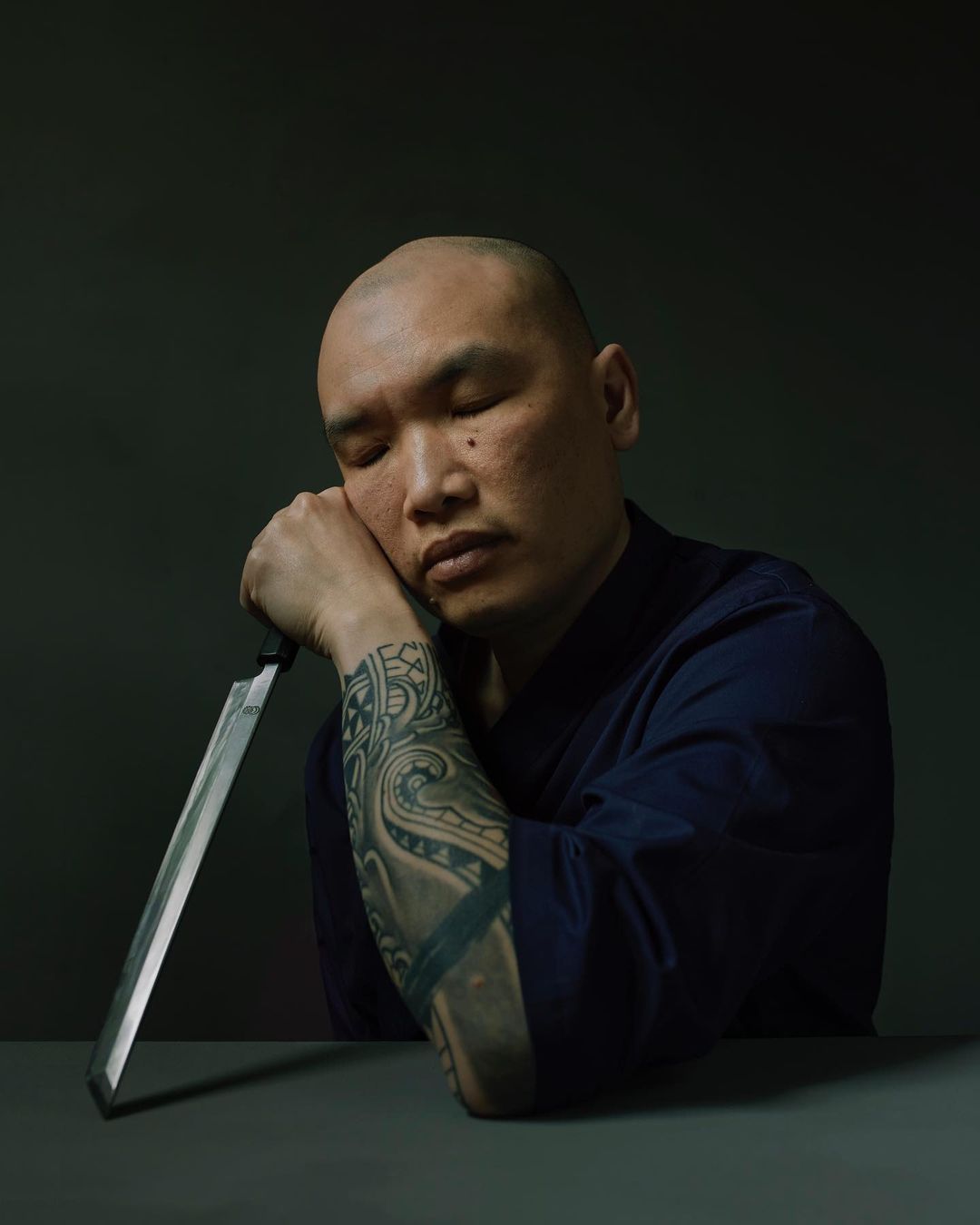 Portrait of a bald adult male with eyes closed resting his head on his tattooed arm which lays on a table next to a knife.