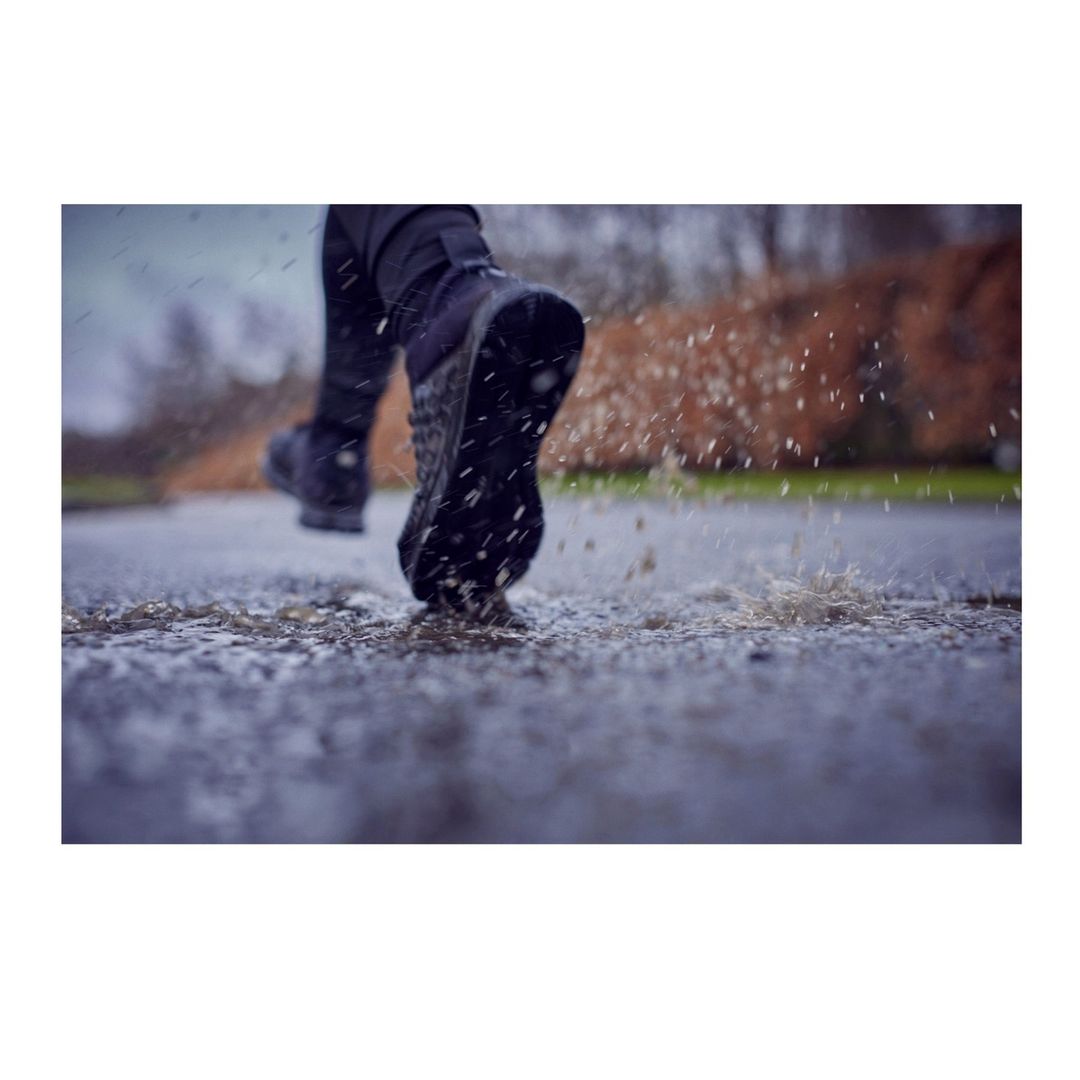 Person in black boots runs through a puddle, kicking up water droplets on a wet surface in an outdoor environment, creating splash.