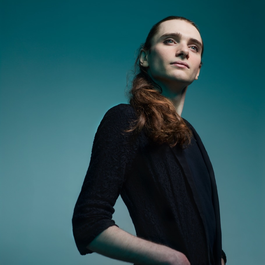 Portrait of a young adult male with long hair, wearing a dark cardigan and shirt, in front of a teal-colored background.