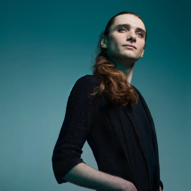 Portrait of a young adult male with long hair, wearing a dark cardigan and shirt, in front of a teal-colored background.