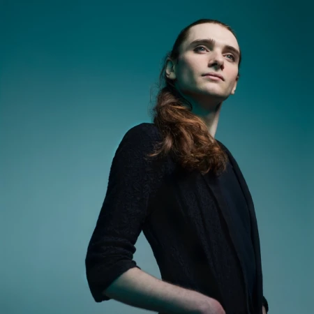 Portrait of a young adult male with long hair, wearing a dark cardigan and shirt, in front of a teal-colored background.