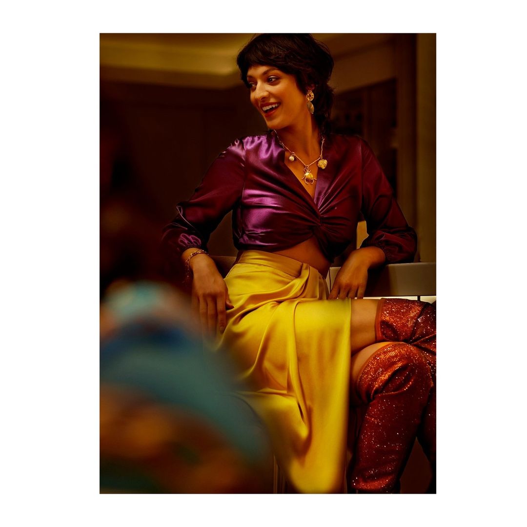 Portrait of a smiling adult woman with dark hair wearing a purple satin shirt, yellow skirt, jewelry and red glitter boots indoors.