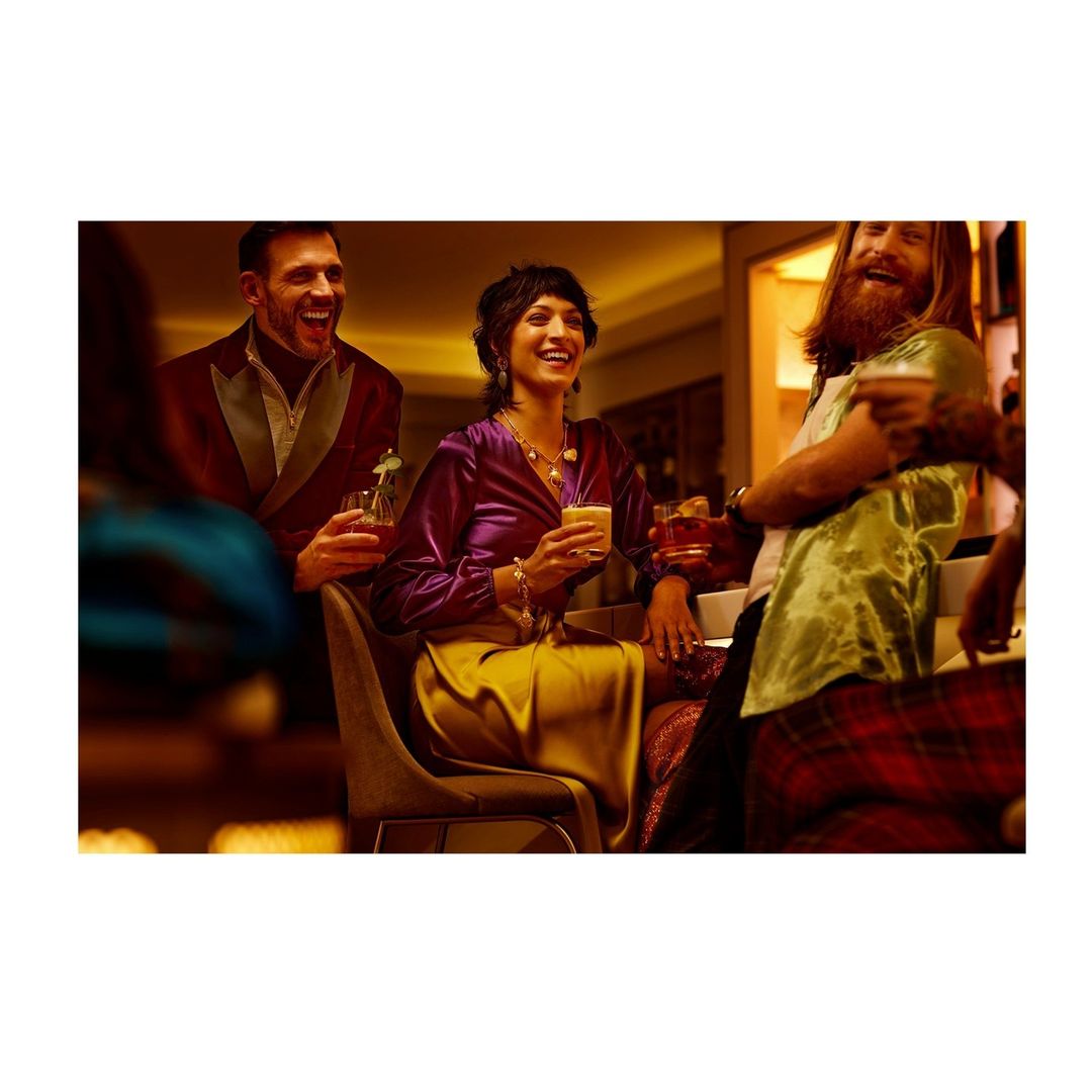 A group of casually elegant people are having drinks and laughing in a warm bar setting. People include men and women with drinks in hand.