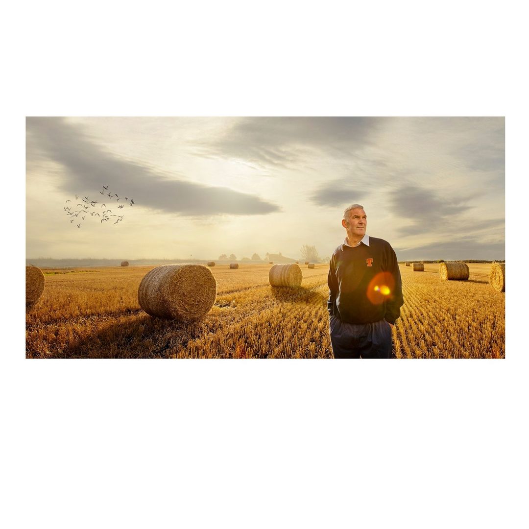 Adult male standing in a harvested field with hay bales, golden sunlight, and cloudy skies adding depth and texture to the rural landscape.