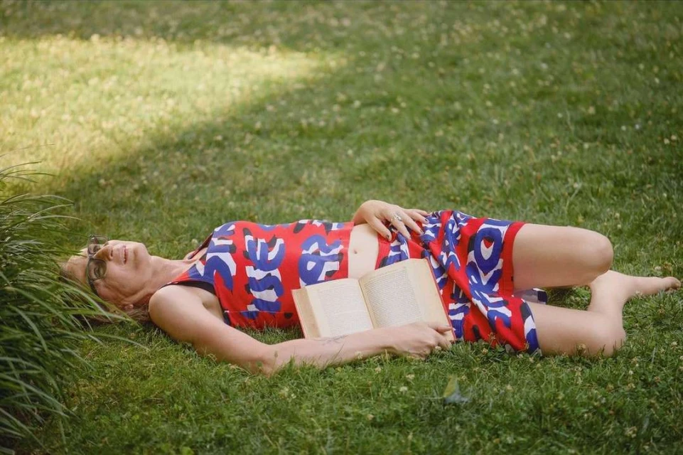 Woman lies on grass reading a book in patterned red and blue dress, barefoot in the sunlight, enjoying the outdoor scene.