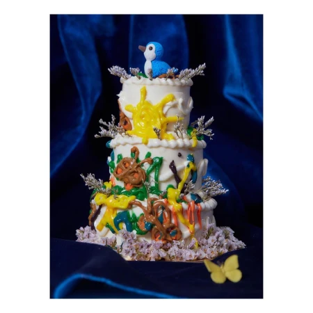 Three-tier white cake decorated with colorful abstract designs and topped with a blue bird figurine set against a dark blue velvet backdrop.