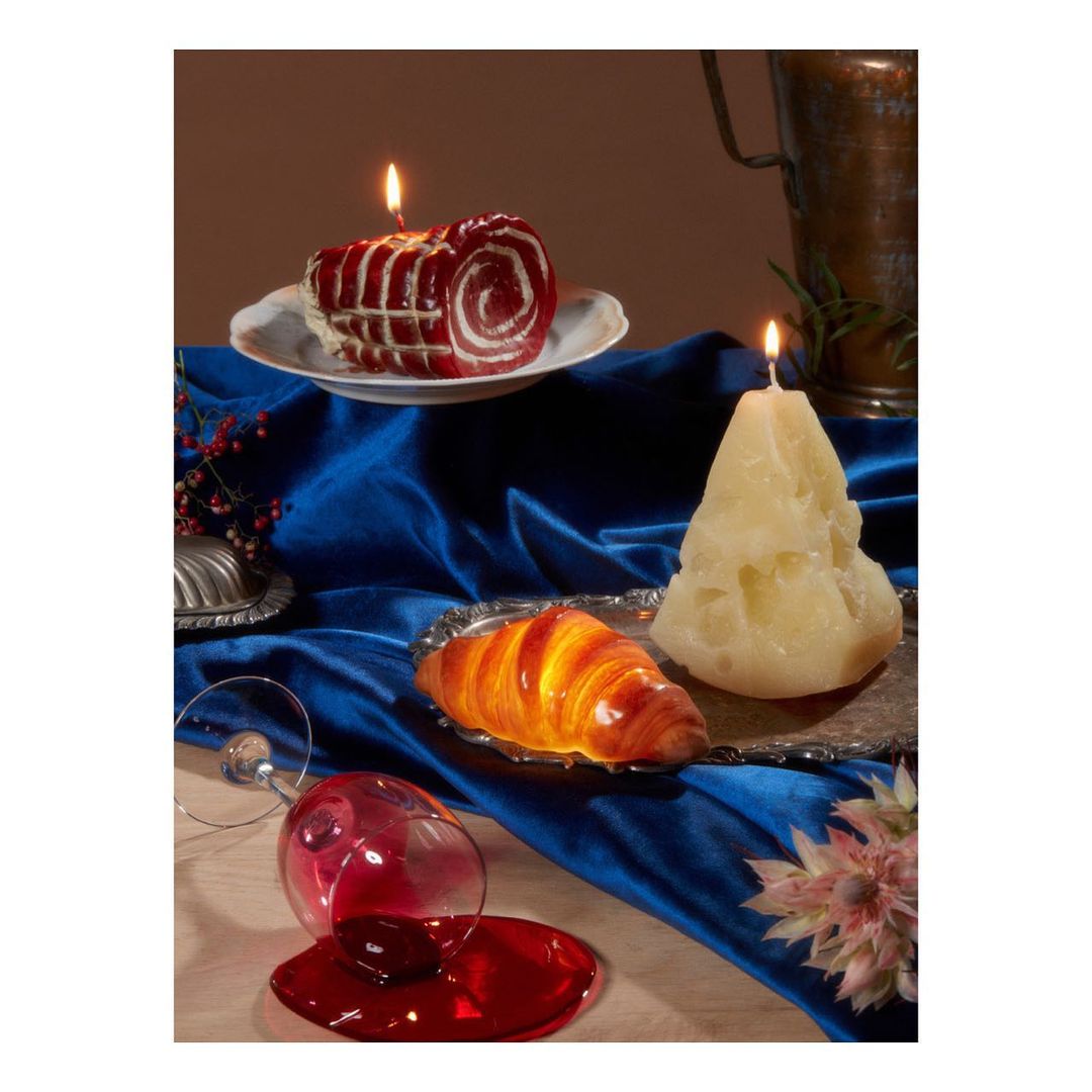 Still life photograph of lit candles shaped like desserts, a croissant, and spilled red wine on a table covered with a blue velvet cloth.