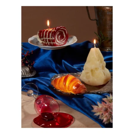 Still life photograph of lit candles shaped like desserts, a croissant, and spilled red wine on a table covered with a blue velvet cloth.