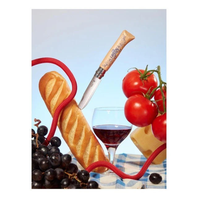 Still life composition featuring bread, wine, grapes, tomatoes, cheese, and a folding knife on a blue and white checkered tablecloth.