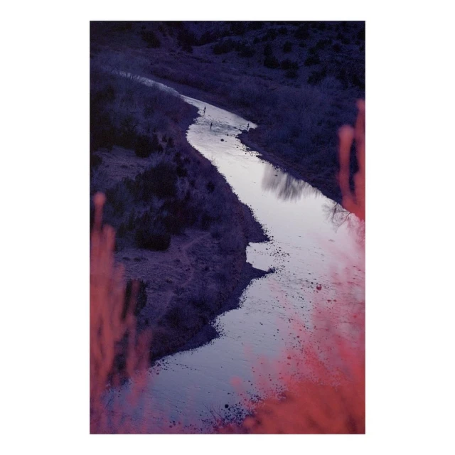 Aerial view of a winding river flanked by hills and vegetation, with figures visible in the distance under a violet sky.