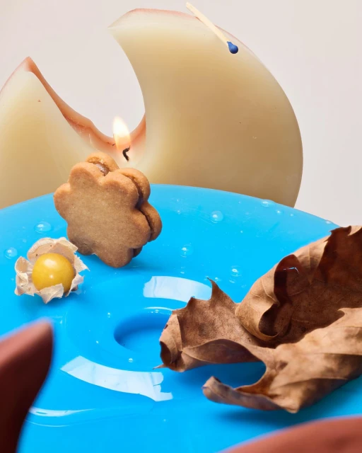 Still life of a cookie candle, a husk cherry, and a dried leaf on a glossy turquoise plate, near a sculpted peach candle.