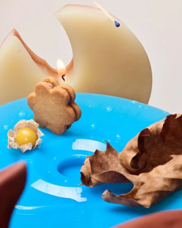 Still life of a cookie candle, a husk cherry, and a dried leaf on a glossy turquoise plate, near a sculpted peach candle.