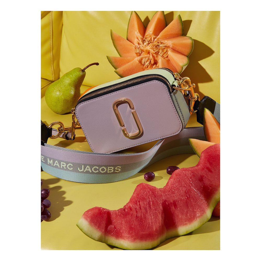 Lilac and mint color-block Marc Jacobs camera bag with melons, grapes, and pear on a bright yellow background in a studio shot.