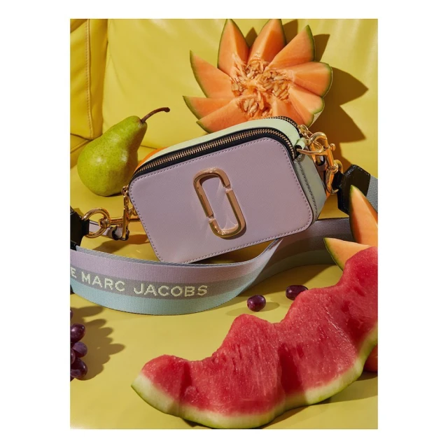 Lilac and mint color-block Marc Jacobs camera bag with melons, grapes, and pear on a bright yellow background in a studio shot.