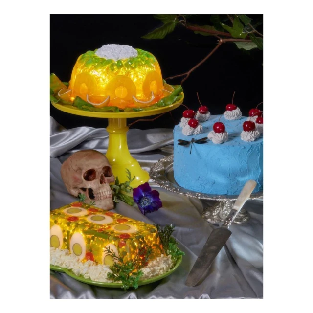 A surreal still life features colorful gelatin molds, a blue frosted cake with cherries, and a human skull on a silver tablecloth.