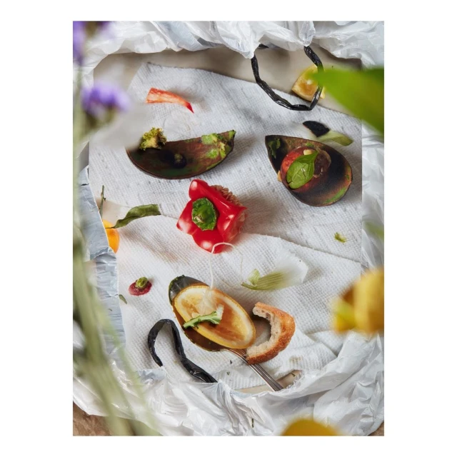 Still life food photography of cooked mussels, sliced bread, and red pepper on a white paper towel and plastic bag.