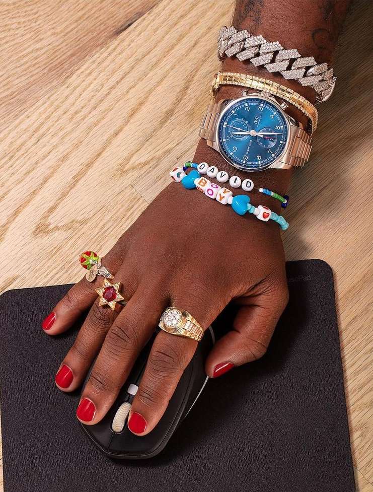 Person wearing gold watch, bracelets, and rings on hand using a black computer mouse on light wood table, manicure is red.
