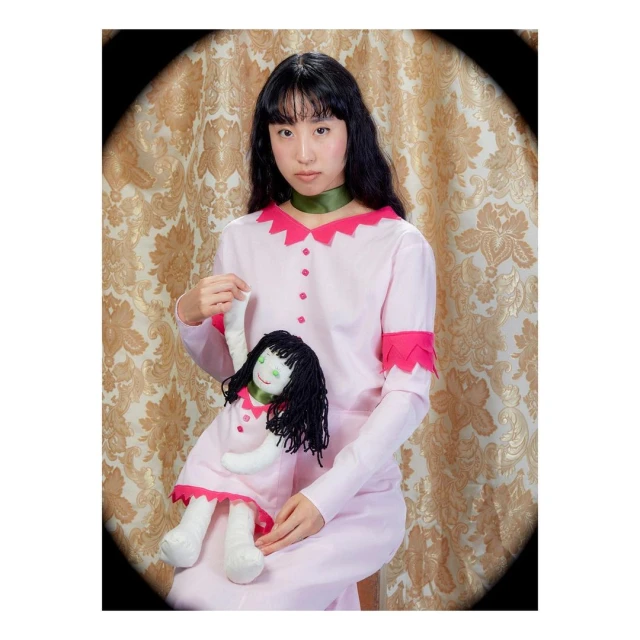 A young woman with black hair in a light pink dress holds a doll in a photo studio with gold damask background, creating a vintage aesthetic.