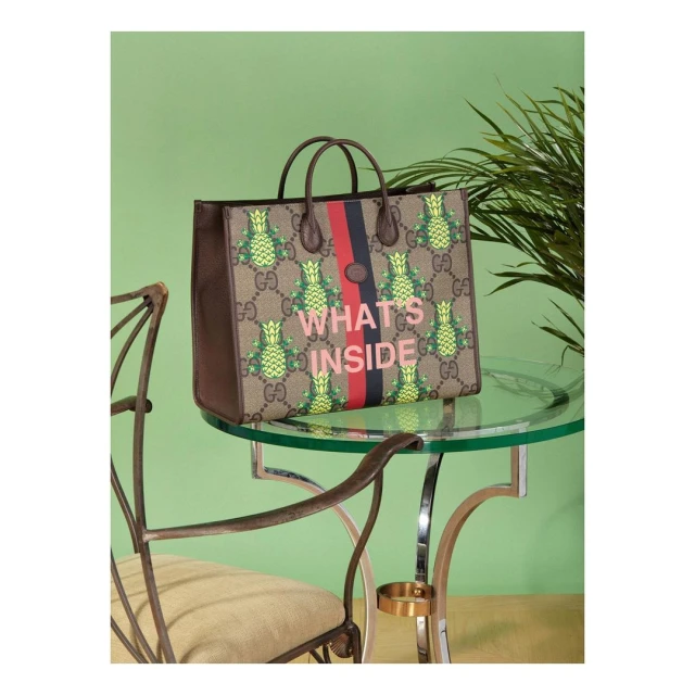Gucci tote bag with pineapple print on a glass-top table next to a metal chair with green wall and tropical plant in background.