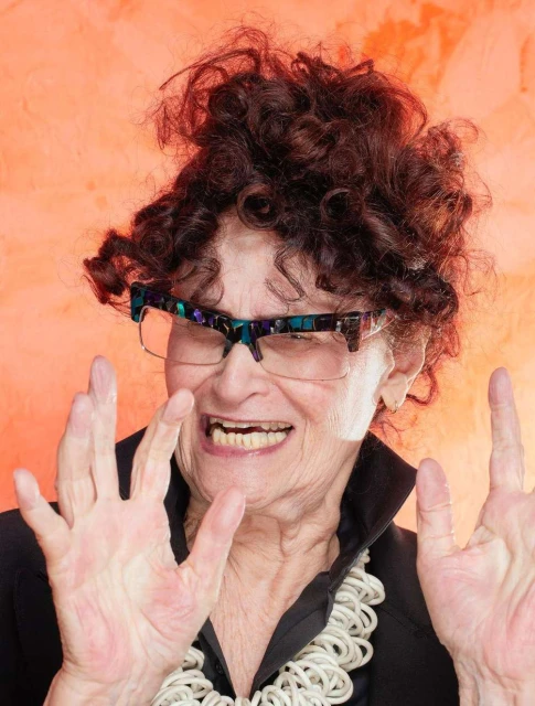 Elderly woman with curly brown hair and glasses smiles with hands raised in a close-up studio portrait against an orange backdrop.