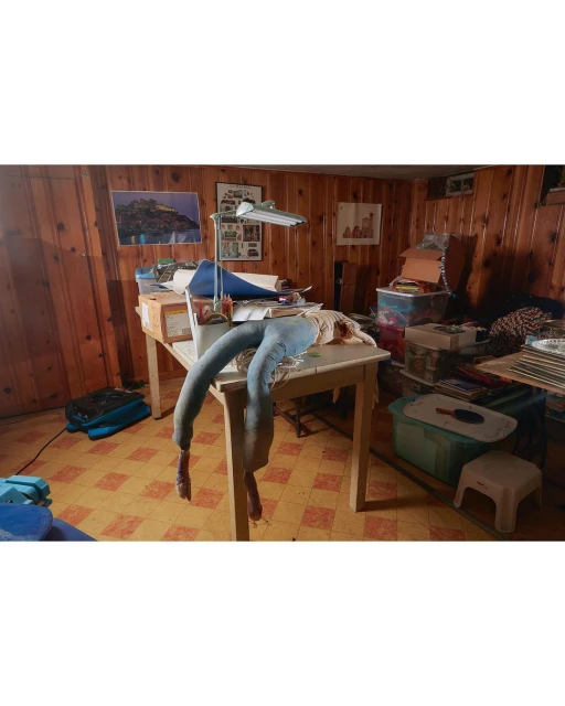 Surreal scene in cluttered room:  table with lamp, jeans with shoes, cardboard boxes, wood paneling, tiled floor, and various objects.