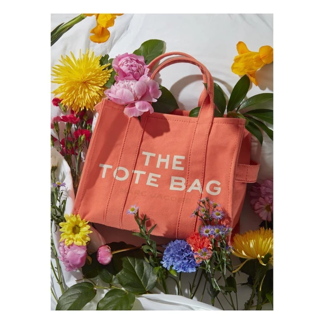 Coral 'The Tote Bag' from Marc Jacobs surrounded by yellow and pink flowers and green foliage, captured indoors against a white cloth.