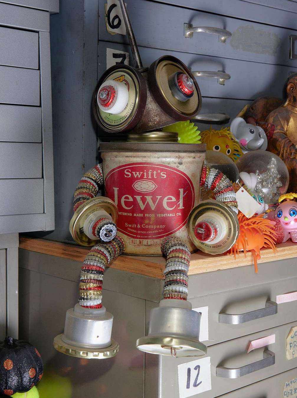 Robot art sculpture made of recycled metal and kitchen container components, resting on a metal cabinet in a room with vintage decor.