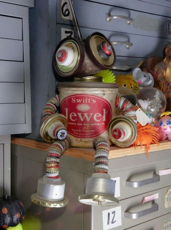 Robot art sculpture made of recycled metal and kitchen container components, resting on a metal cabinet in a room with vintage decor.