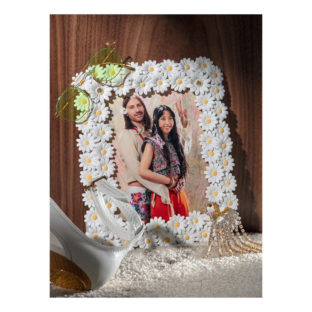 Daisy-framed photo of a man and woman in vintage attire with white high heels, daisy sunglasses, and gold jewelry on a shag rug.