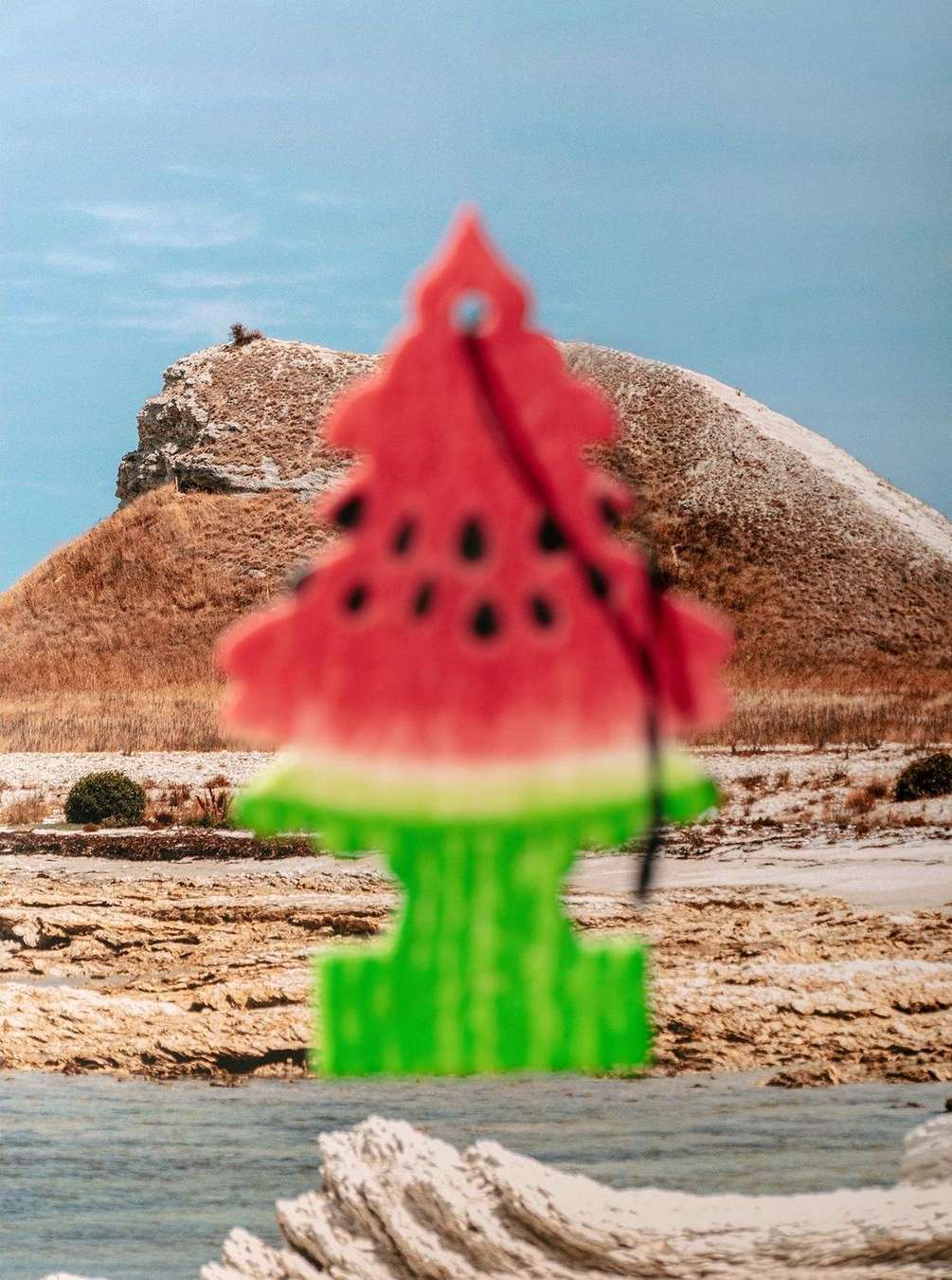 Whimsical watermelon-shaped car air freshener against a backdrop of mountains and water, creating a playful, summery scene.