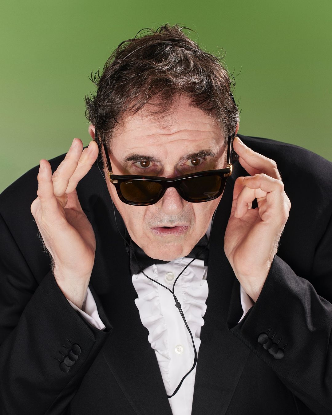 Man in tuxedo and sunglasses poses with hands near face against green backdrop, creating a curious and comedic visual portrait.