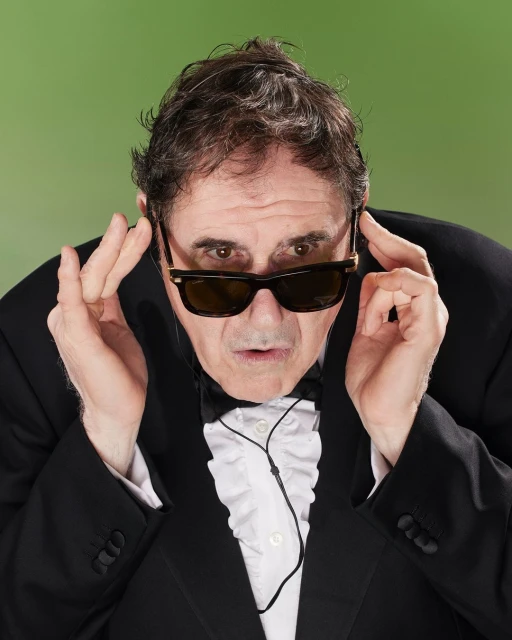 Man in tuxedo and sunglasses poses with hands near face against green backdrop, creating a curious and comedic visual portrait.