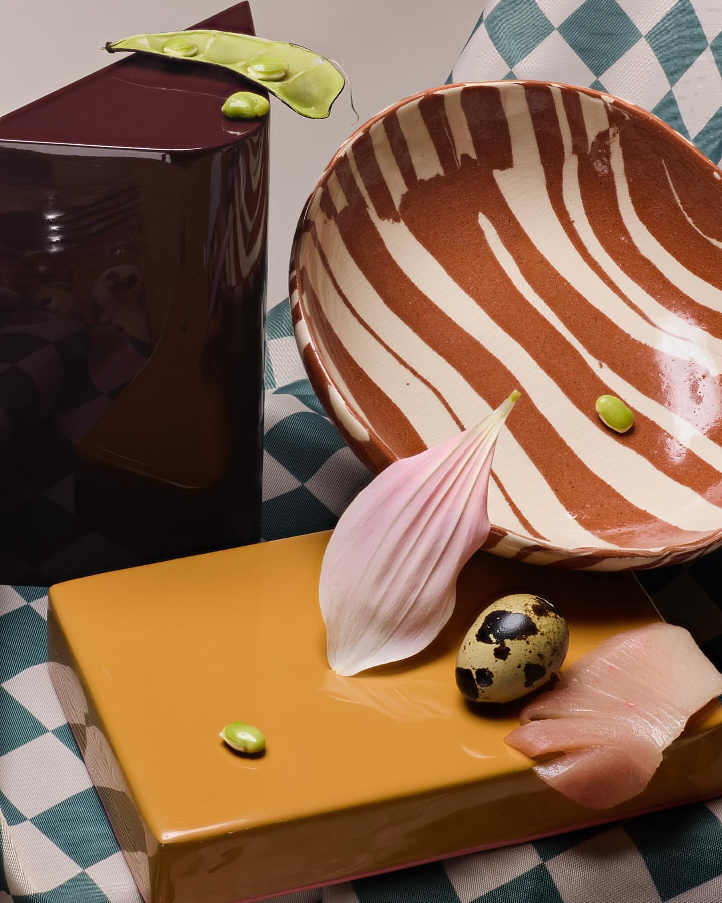 Still life composition featuring a brown ceramic vase, striped bowl, tan platform, with a flower petal, speckled quail egg and green beans.