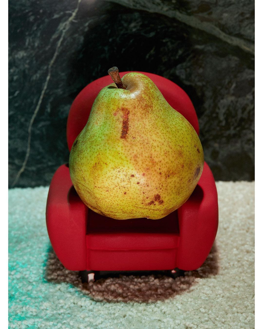 A vibrant pear rests on a miniature red chair against a dark marble backdrop, creating a playful food still life.