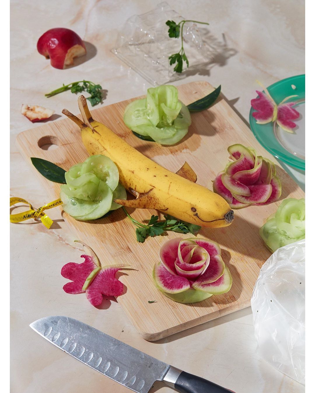 Banana carved as a dolphin on a wooden cutting board with flowers made from cucumber and radish, apple, knife, marble surface.