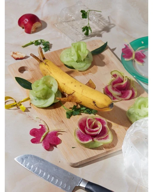Banana carved as a dolphin on a wooden cutting board with flowers made from cucumber and radish, apple, knife, marble surface.