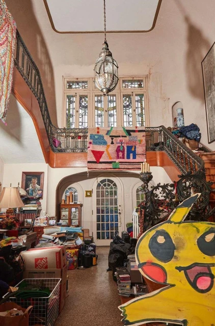Interior of a cluttered foyer with ornate staircase, windows, and decorative objects, featuring a whimsical Pikachu cutout and assorted boxes.