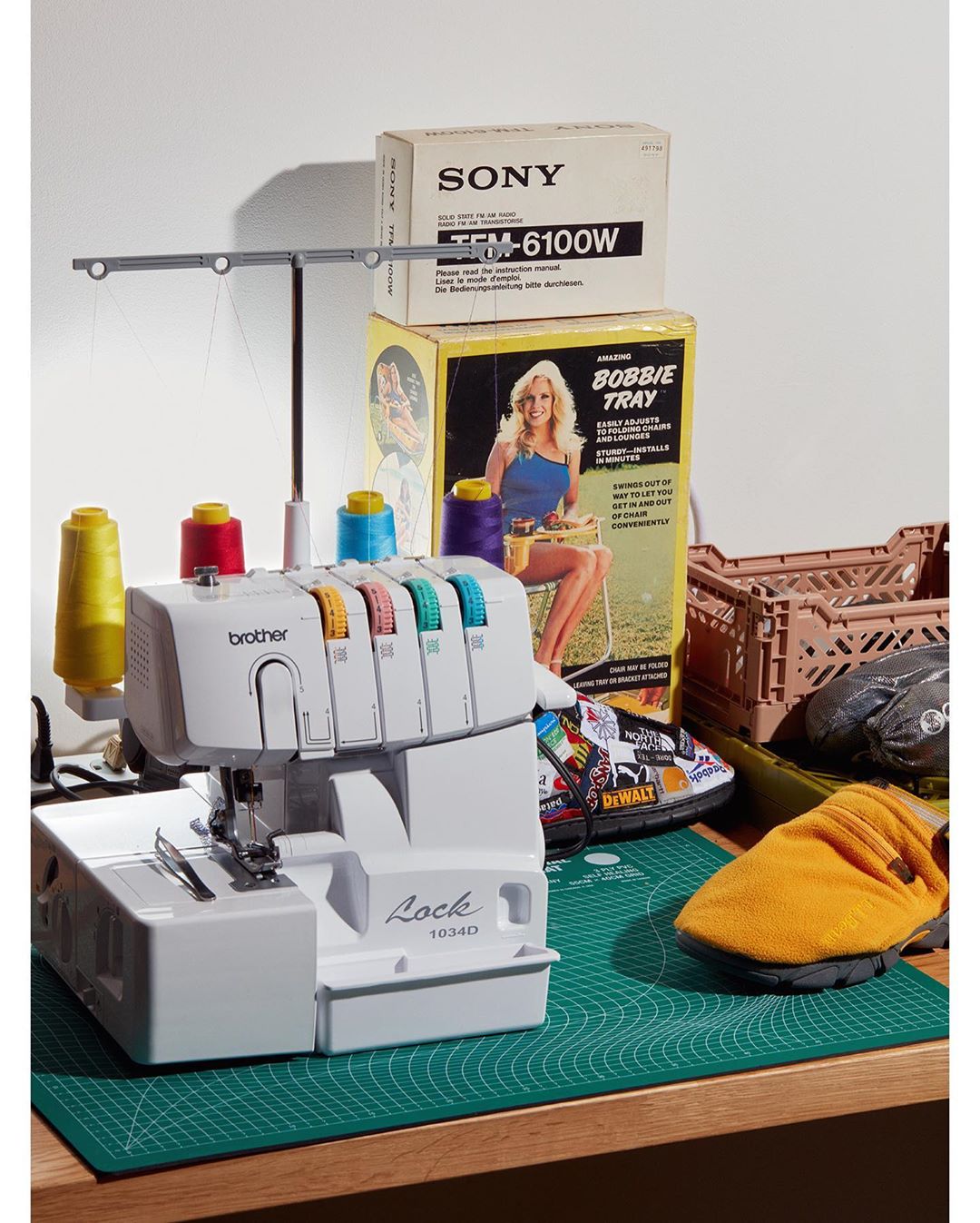 A white Brother sewing machine rests on a green cutting mat, surrounded by spools of thread, vintage boxes, and a pair of mustard slippers.