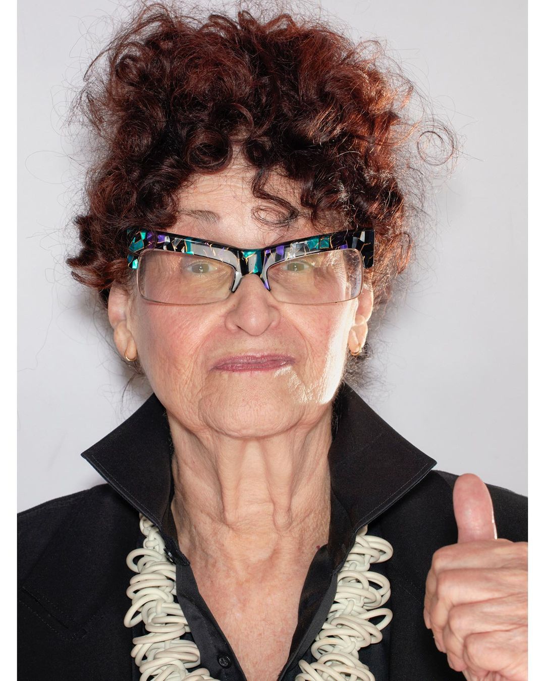 Elderly woman in glasses and black blazer gives a thumbs-up in a studio portrait with a white necklace and curly red hair.