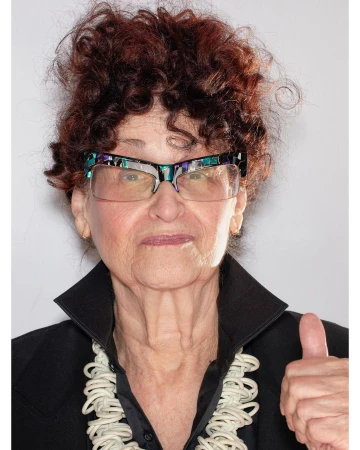 Elderly woman in glasses and black blazer gives a thumbs-up in a studio portrait with a white necklace and curly red hair.