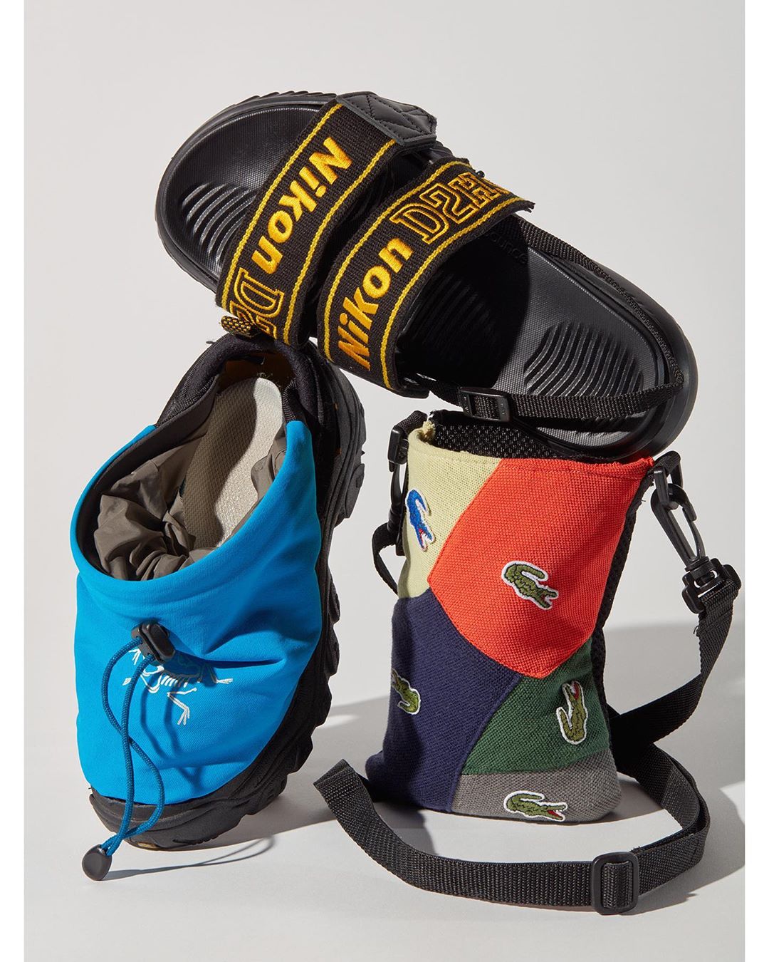 Still life shot showcasing a black sandal with 'Nikon D2H' straps, alongside a blue pouch and a Lacoste-themed sling bag, all on white backdrop.