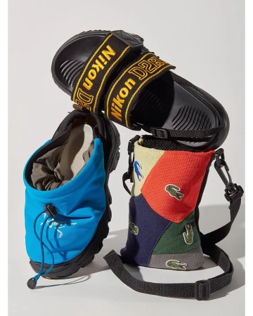 Still life shot showcasing a black sandal with 'Nikon D2H' straps, alongside a blue pouch and a Lacoste-themed sling bag, all on white backdrop.