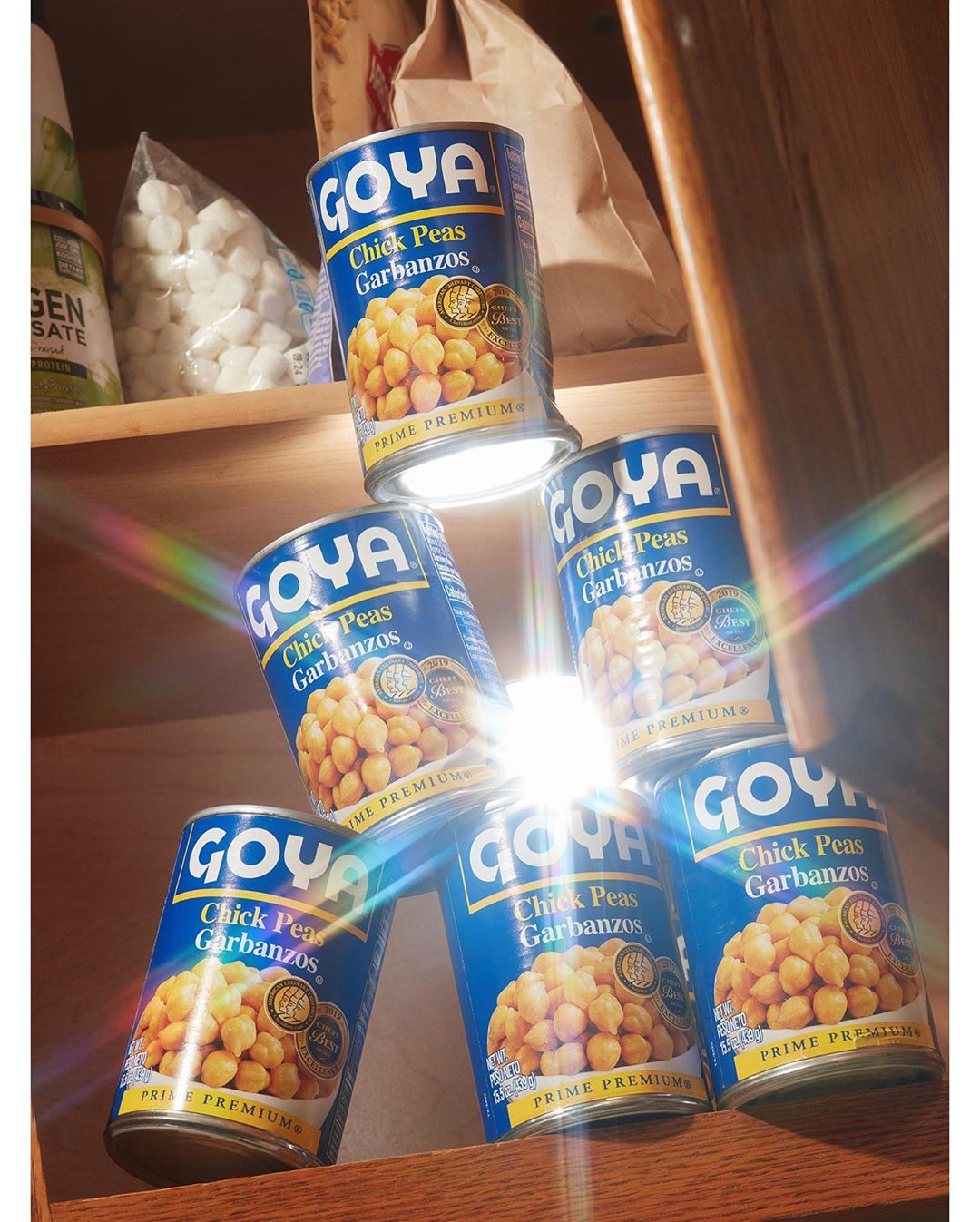 Stack of Goya Chick Peas Garbanzos cans arranged pyramid-style on a shelf. Food photography in a pantry or kitchen setting with ample product visibility.