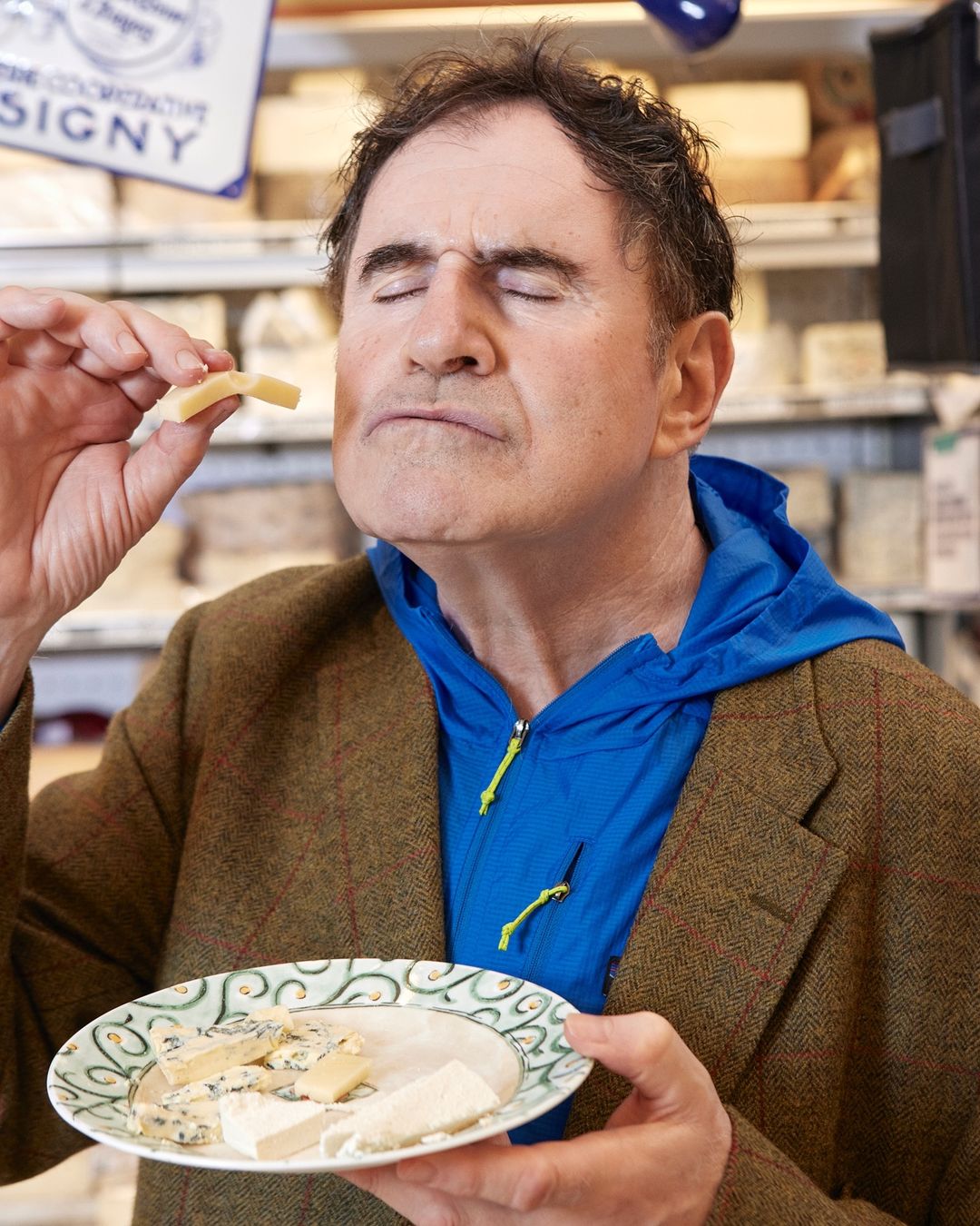 Man with eyes closed holding cheese slice and a plate of assorted cheeses in a deli, wearing a blue jacket and tweed coat.