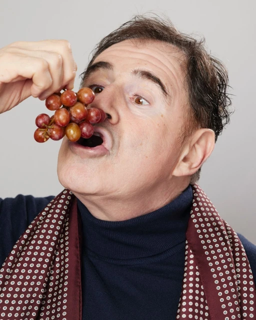 Adult male holding grapes with open mouth, brown hair, wearing a turtleneck, indoors in front of plain white background, close-up shot.