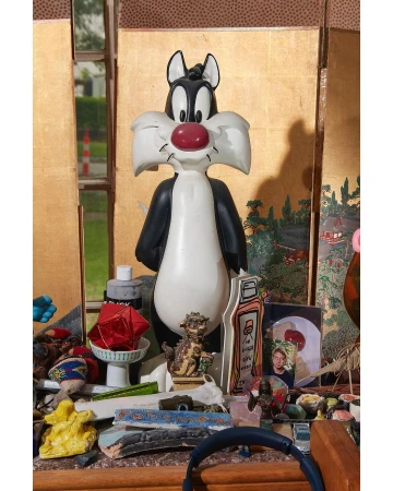 Sylvester the Cat statue in front of a gold screen, surrounded by assorted objects on a wooden surface. A cartoon-themed indoor scene.