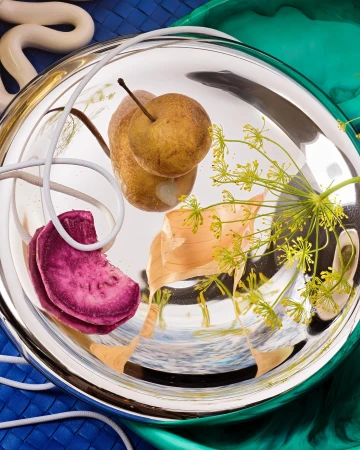 Overhead shot of a pear, onion, beetroot and flower on a reflective plate, set against a backdrop of blue and green cloth.