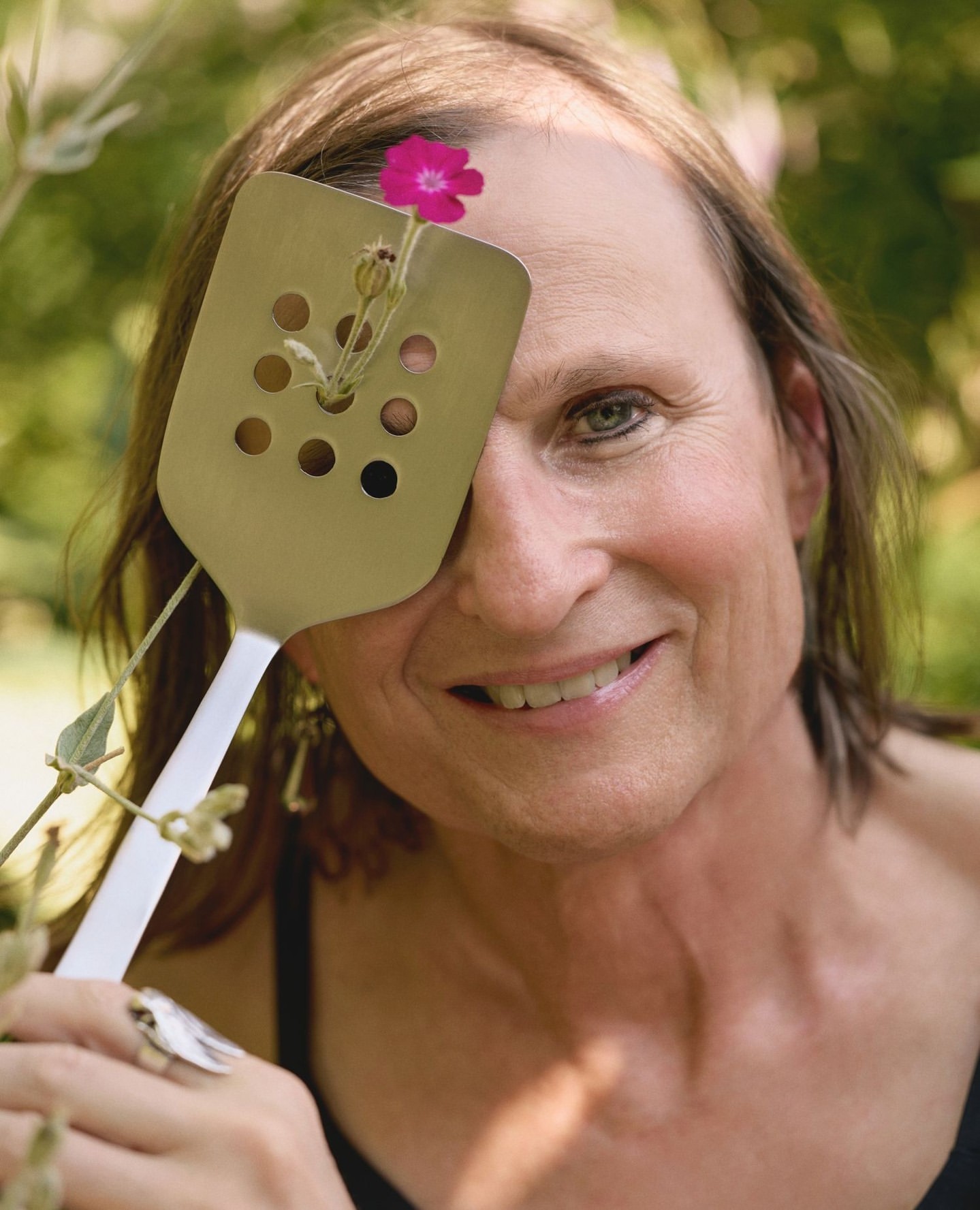 Portrait of a person holding a slotted metal spatula with a small pink flower poking through a hole in front of their eye.