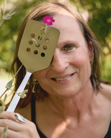 Portrait of a person holding a slotted metal spatula with a small pink flower poking through a hole in front of their eye.