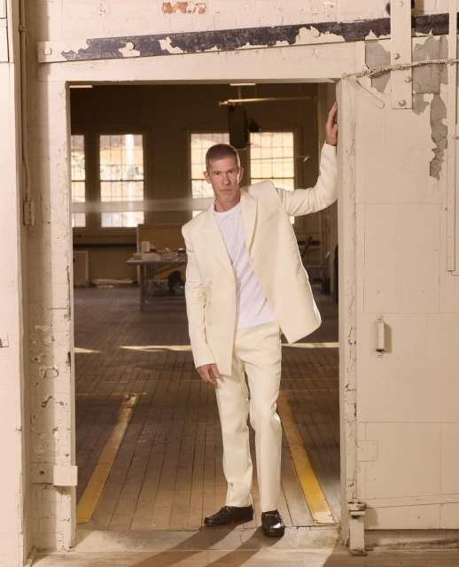 Adult male in off-white suit and black dress shoes stands in an industrial doorway with peeling paint and weathered wooden floors.